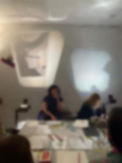 Four people sit around a table, on top of which there is printed material and pens and pencils. There two overhead projectors, one in each side of the table, projecting images to a wall. One of the people is writing on a sheet of paper, whose image is being projected on the wall.