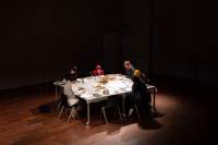 Six people sit around a rectangular table. There is a spotlight shining on the table; the space around it is in the dark.