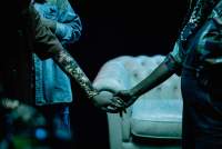 Close-up of two people holding hands. A sofa appears in the background.