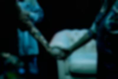 Close-up of two people holding hands. A sofa appears in the background.