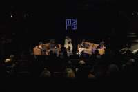 An audience sits in an auditorium. On stage, five people sit in two sofas and a chair, having a discussion. The Melkweg logo is being projected above them.