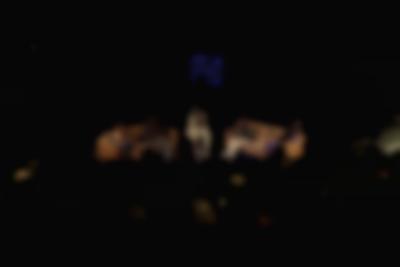 An audience sits in an auditorium. On stage, five people sit in two sofas and a chair, having a discussion. The Melkweg logo is being projected above them.