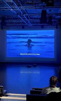 A film is being projected in a cinema room. On screen, someone is submerged up to their chest in the ocean. A subtitle reads: 