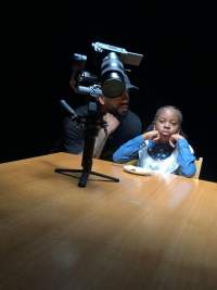 A child and an adult person sit at a table. In front of them, a camera on a gimbal has been left on the table.