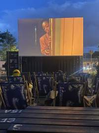 Several deck chairs face a large outdoor screen, onto which a film is being projected. On screen, a feminine-presenting person rests their face on a door frame.